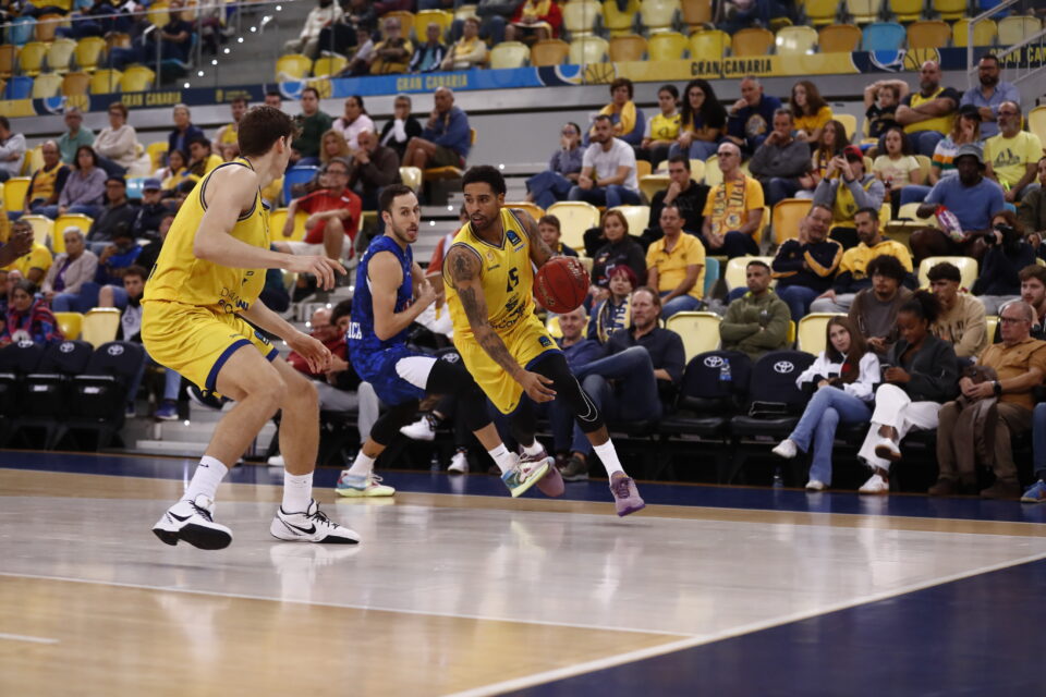 Jugadores de baloncesto en acción durante un partido en el estadio, con público animando.