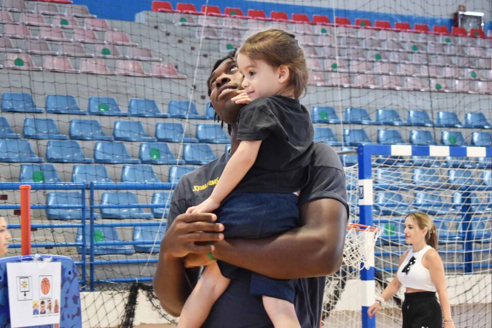 Entrenador sostiene a una niña mientras ambos sonríen en el polideportivo.